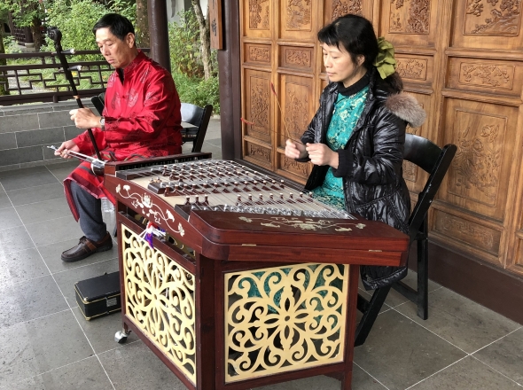 Two Chinese musicians playing traditional instruments in front of a adorned wooden panels  